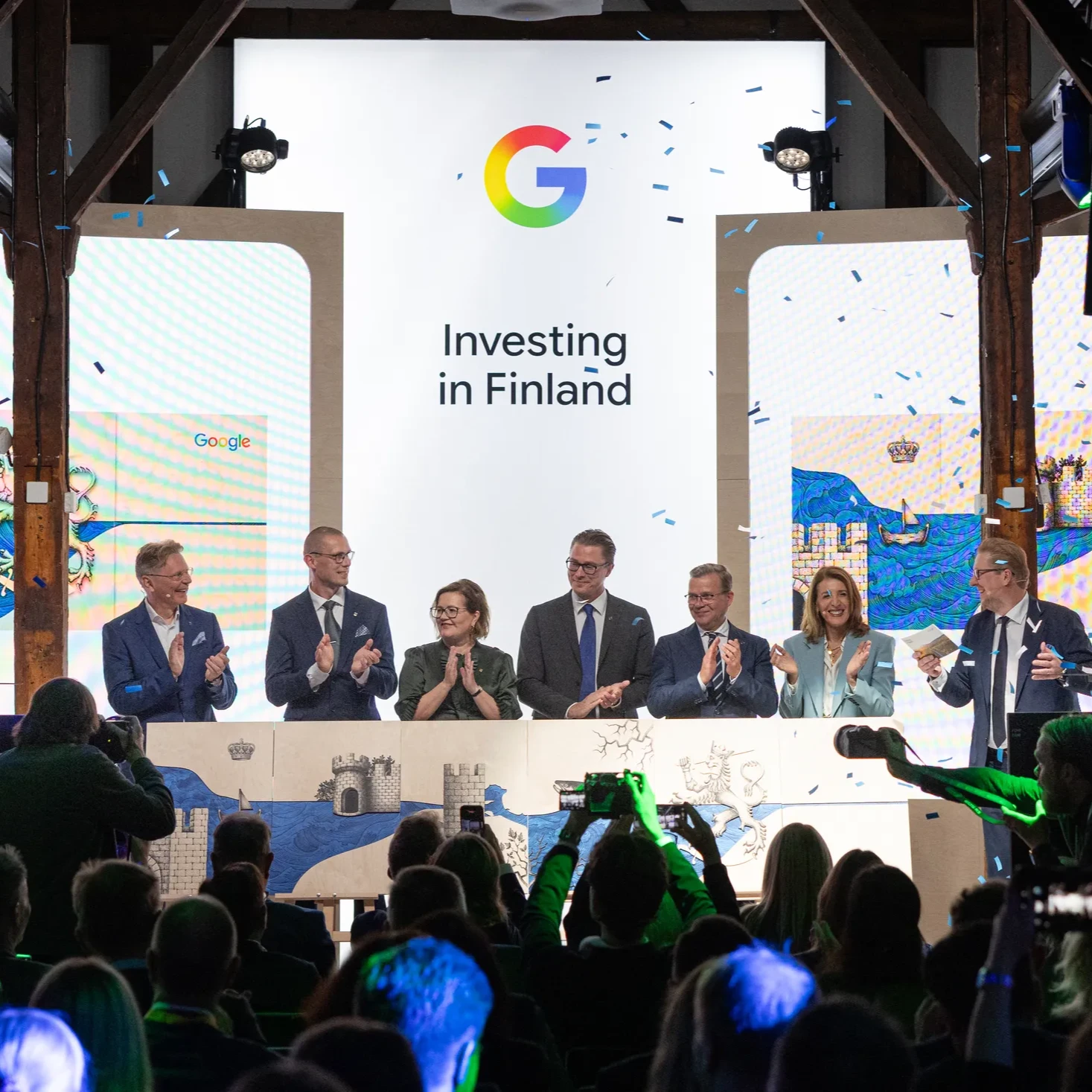 Seven people standing on a stage in front of a piece of art and screens that read Investing in Finland and blue and white confetti