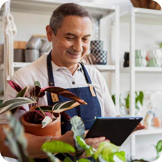 A smiling man wearing a denim apron holds a potted houseplant in one arm while checking a black tablet screen in a plant shop.