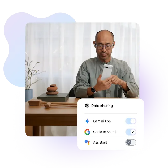 A man with glasses and earbuds sits at a wooden desk, focused on the white smartphone in his hands. He is wearing a grey long-sleeved shirt and a smartwatch. On the desk to his left, there is a small stack of balancing stones and two wooden mallets. An overlaying menu titled "Data sharing" shows toggle switches for the Gemini App and Circle to Search turned on, while the Assistant toggle is turned off.