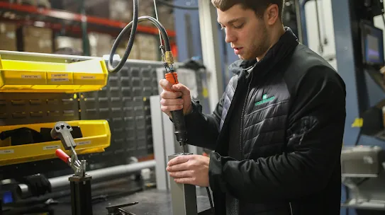 A young man in a black work jacket with the "Kesseböhmer" logo uses a high-torque pneumatic screwdriver to assemble a metal component in a brightly lit industrial warehouse. Yellow storage bins are visible in the background, representing the type of high-skilled technical labor addressed by bipartisan AI workforce legislation.