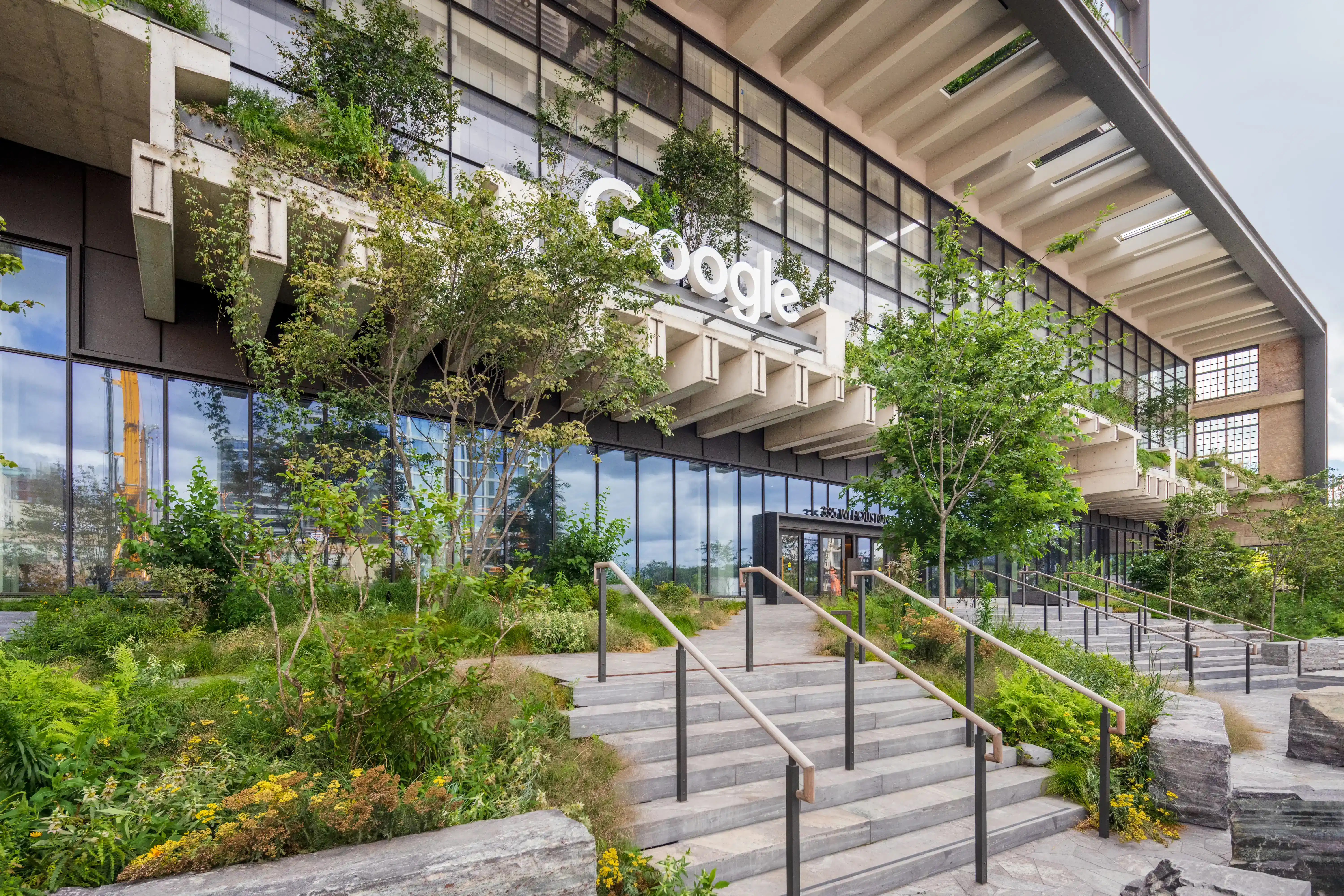 The modern exterior of a Google building features large glass windows, lush greenery growing on ledges, and a prominent "Google" sign above a set of concrete steps leading to an entrance.