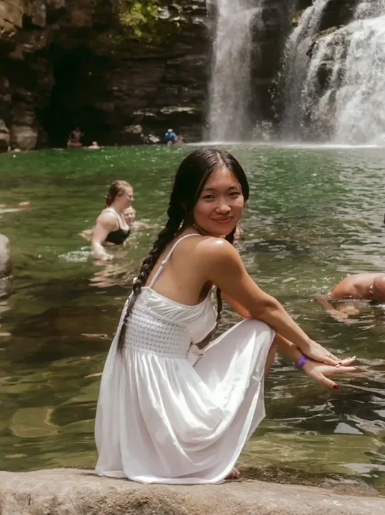 A person gets their photo taken wearing a white summery dress sits on a rock with waterfalls and other people in the background. A prompt asks Gemini to remove the people in the background. The people disappear from the photo with Gemini confirming the task is complete.