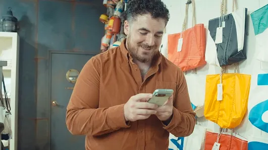A smiling man in a rust-colored shirt looking at his smartphone inside a store with colorful tote bags hanging on the wall.