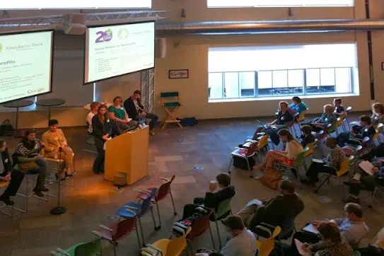 Panel speakers and a presenter at a wooden podium speaking to an audience seated in a bright conference space with presentation slides displayed on overhead screens