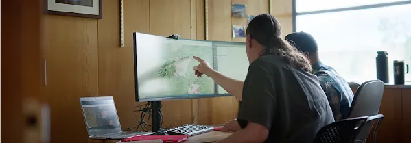 Two people sit side-by-side at a desk looking at a map on a computer monitor with the person on the left pointing to the screen
