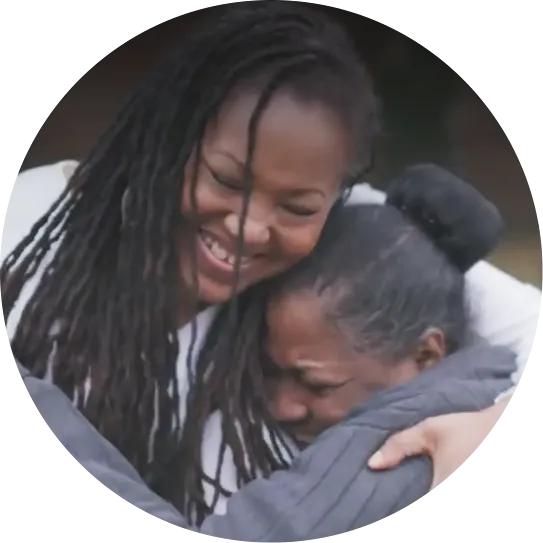 A circular cropped portrait of a smiling woman warmly embracing another woman in a supportive hug.