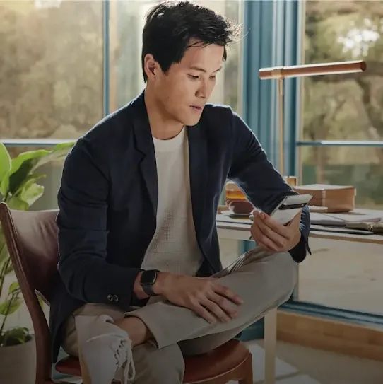 A man sits in a leather chair with his legs crossed, looking at a smartphone in his hand. He is wearing a dark blue blazer over a white shirt, light-colored trousers, white sneakers, and a smartwatch. In the background, there is a desk with a lamp and a large window looking out onto greenery.