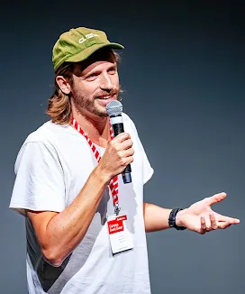 A host addresses the audience from the stage while wearing a cap.