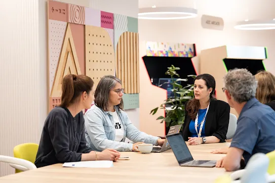 Cinq personnes sont assises autour d'une table dans un bureau moderne et discutent. Deux personnes ont un ordinateur portable devant elles. En arrière-plan, on distingue des décorations murales colorées et des bornes d’arcade. Ces personnes se trouvent dans l'Accessibility Discovery Center de Google Suisse.