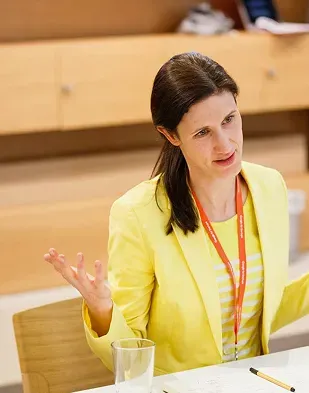 A woman in a yellow suit is speaking and moving her hands expressively.