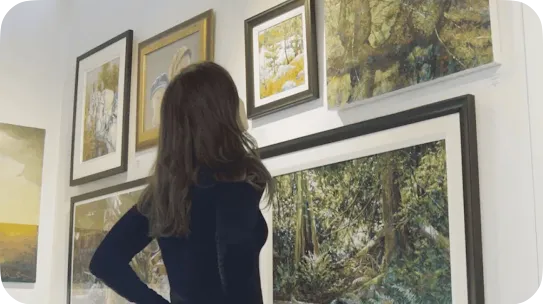 A woman with long dark hair standing with her hands on her hips, admiring various framed nature and landscape oil paintings displayed on a white gallery wall.
