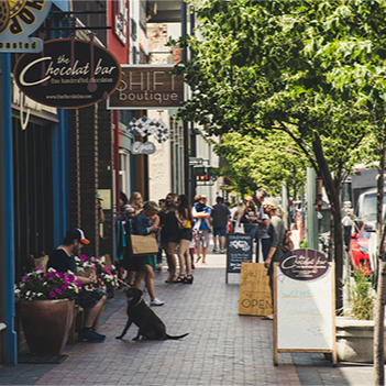 A man in a baseball cap sits on the edge of a large planter filled with flowers on a busy city sidewalk