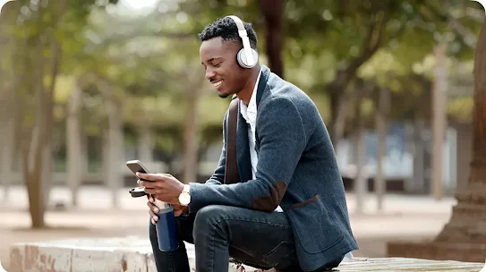 A professional, Black man wearing over-ear headphones is smiling while holding his mobile device and a water bottle while sitting outside on a park bench.