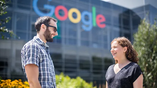Two people, a man with a beard and sunglasses and a woman with wavy hair, stand outside a modern glass building with a large, colorful Google logo. They are smiling and facing each other, with a garden of yellow flowers in the foreground.
