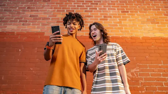 Zwei junge Erwachsene halten Android Smartphones in der Hand und machen ein Selfie vor einer roten Backsteinwand – moderne Handy-Nutzung im Alltag.