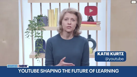 A screenshot of a video broadcast featuring Katie Kurtz from YouTube speaking to the camera. She is a woman with blonde, shoulder-length hair wearing a dark blue crewneck sweater. A lower-third graphic on the right displays her name, "KATIE KURTZ," and handle, "@youtube." The bottom news banner reads "YOUTUBE SHAPING THE FUTURE OF LEARNING" next to a "CHICAGO NOW" logo. The background features a modern wooden shelf decorated with yellow books, a trailing green plant, a circular blue vase, and a prominent 3D YouTube play button logo.
