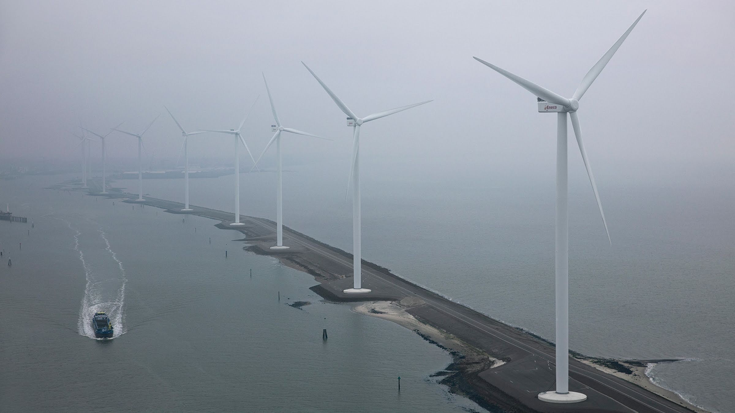 Wind turbines near water, standing on along a thin strip of land in foggy weather.
