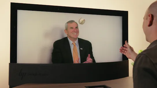 A man wearing a blazer and yellow tie is tossing a ball up into the air on a Google Beam screen. This is a static image for the Google Beam 3D video communication system, highlighting its ability to convey subtle interactions and handle objects with a realistic sense of depth.