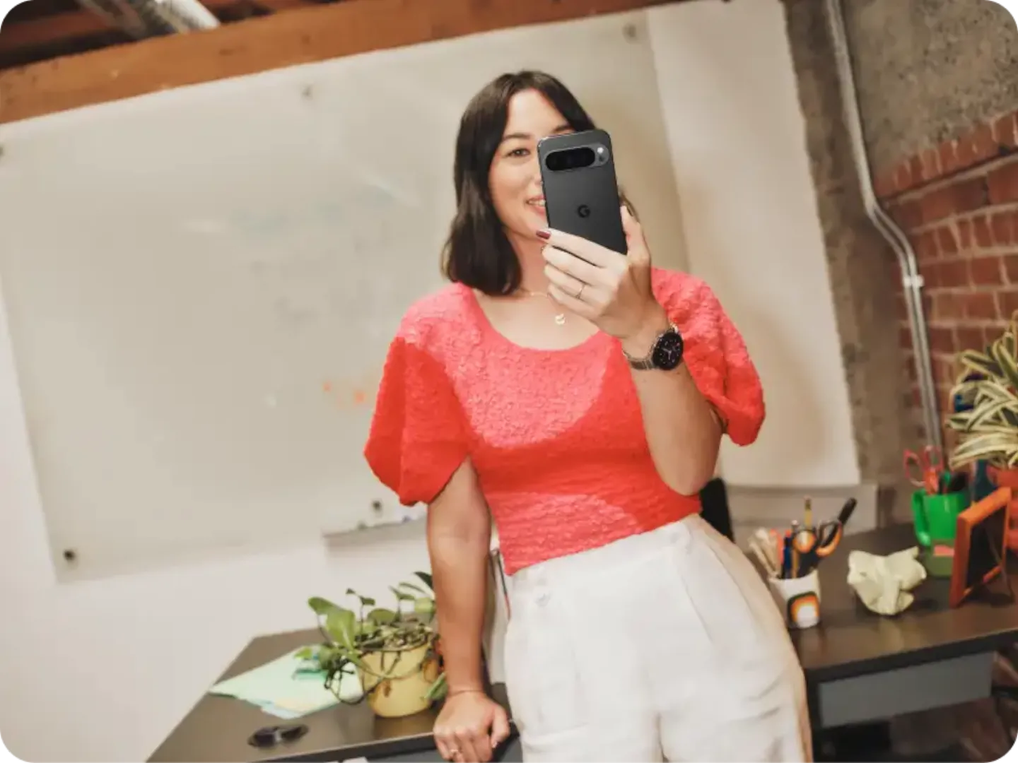 Woman in a bright pink top is holding a pixel 9 in her hand while leaning on her desk.