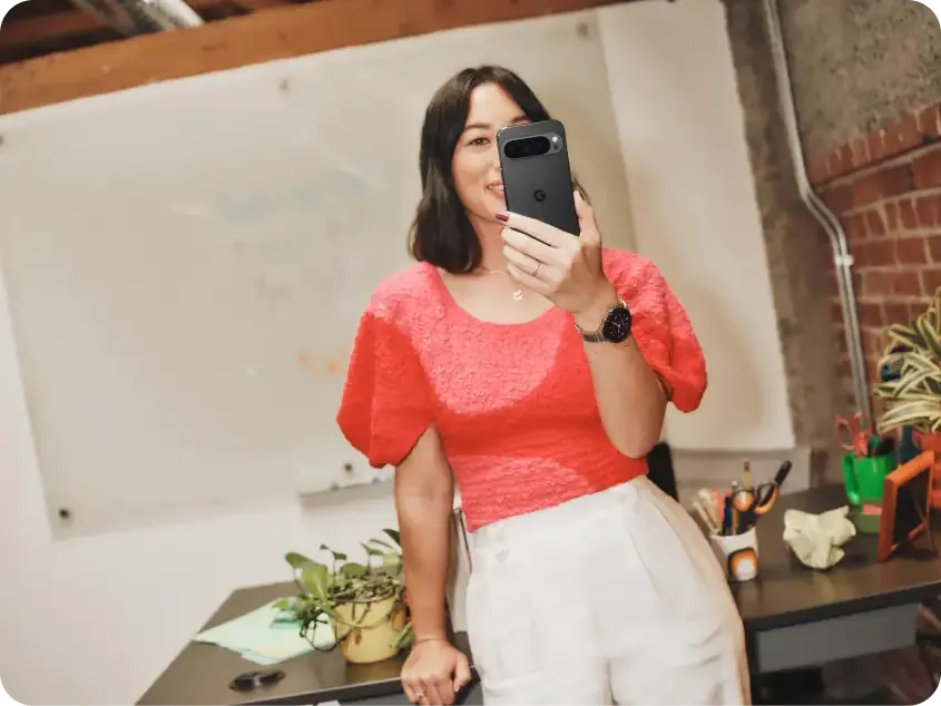 Woman in a bright pink top is holding a pixel 9 in her hand while leaning on her desk.