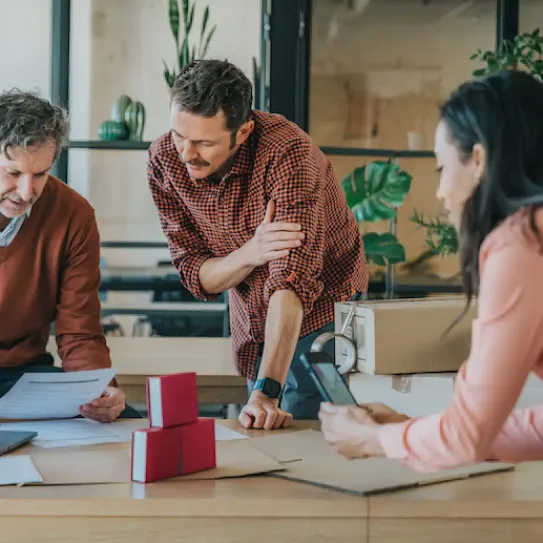 Two men review a document together at a wooden table while a woman holds a smartphone nearby; cardboard boxes and packing tape sit on the table.