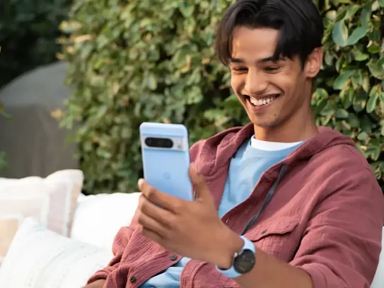 A man sitting on a patio outdoors while looking at a smartphone and smiling.