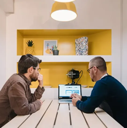 Deux hommes de dos assis de part et d’autre d’une table, avec un ordinateur portable au milieu qu’ils regardent tous les deux