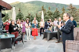 A man in a dark suit speaks into a microphone, addressing a diverse group of attendees at an outdoor evening reception. The event is held on a stone patio with modern lounge furniture, surrounded by evergreen trees and a scenic mountain backdrop under a soft twilight sky.