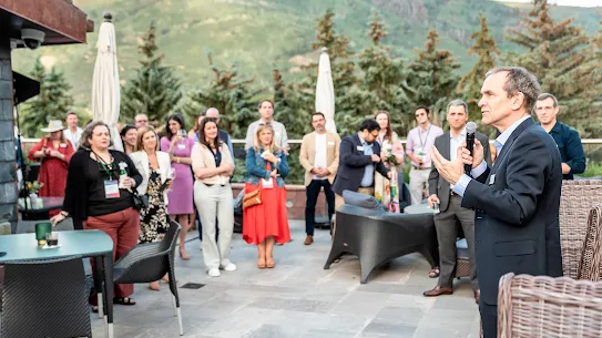 A man in a dark suit speaks into a microphone, addressing a diverse group of attendees at an outdoor evening reception. The event is held on a stone patio with modern lounge furniture, surrounded by evergreen trees and a scenic mountain backdrop under a soft twilight sky.
