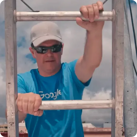 A man wearing a blue Google t-shirt, sunglasses, and a grey baseball cap climbs a metal ladder outdoors against a cloudy sky background.
