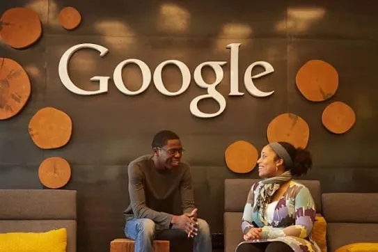 Two people sitting on modern lounge chairs having a conversation in front of a wall featuring a large Google logo surrounded by round wooden accents