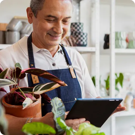 A smiling man wearing a denim apron holds a potted houseplant in one arm while checking a black tablet screen in a plant shop.