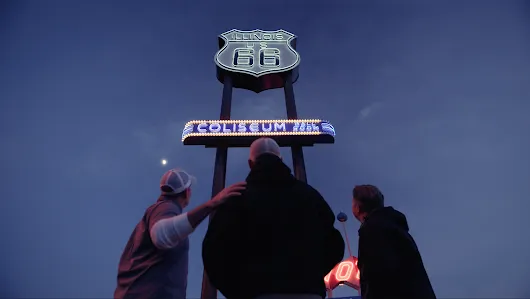 Three people look up at a lighted road sign against the night sky 