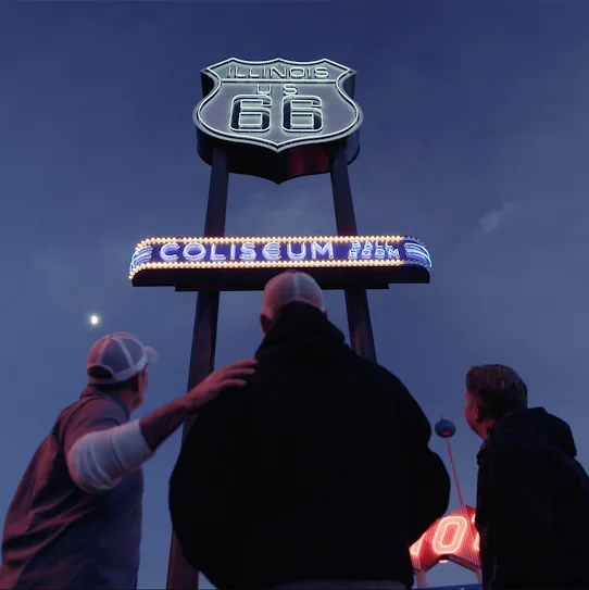 Three people look up at a lighted road sign against the night sky 