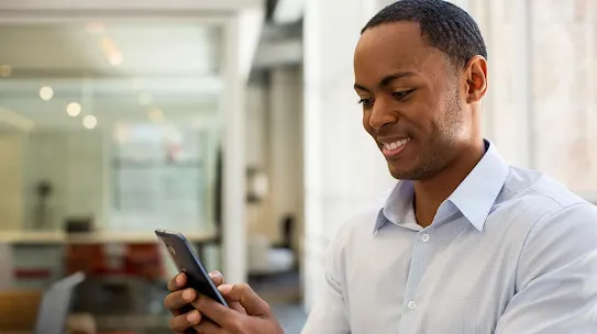 A smiling man in a light blue shirt using a smartphone in a modern, brightly lit office environment.