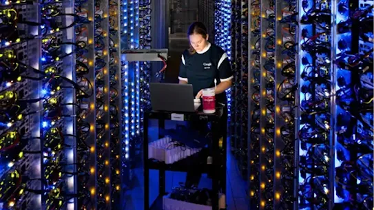 A woman wearing a Google t-shirt working on a laptop on a cart in a data center aisle, surrounded by server racks with blue and orange lights.