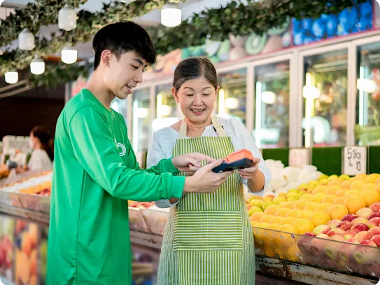 A young man in a green shirt showing a female fruit vendor how to use a handheld credit card terminal in a market.