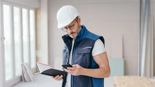 A construction worker wearing a white hard hat and safety vest, holding a notebook while checking his smartphone on a job site.