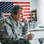 A soldier in camouflage uniform wearing a headset and writing on paper at a desk with an American flag in the background.