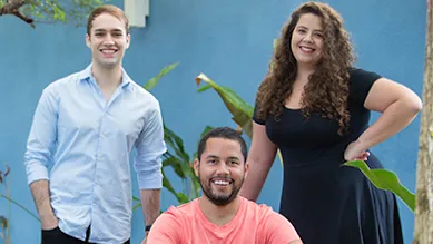 Cuidas founders João, Matheus, and Deborah pose together in front of a blue wall.