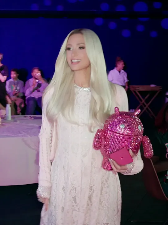 Paris Hilton in a pink lace dress holding a bedazzled pink Android mascot, smiling and waving as she walks past a table.