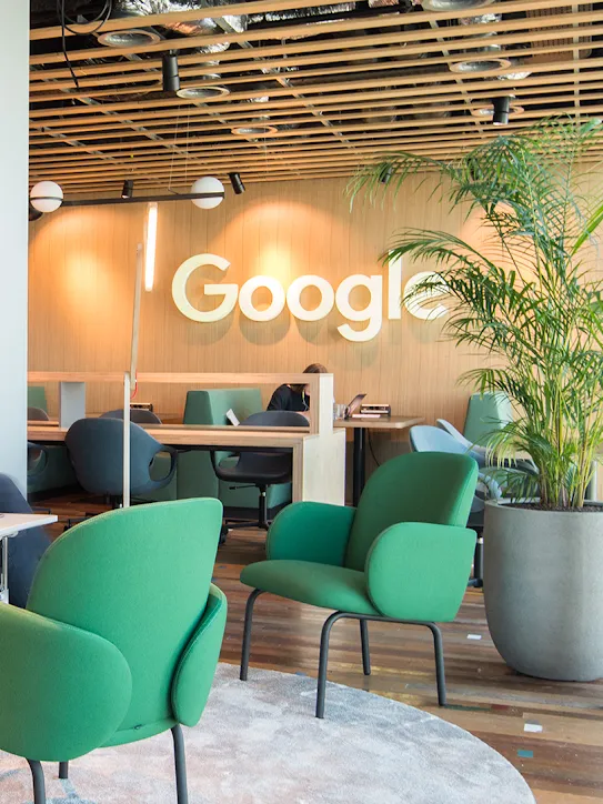 ワークスペースで 2 人が座っており、背景には Google ロゴが掲げられている。