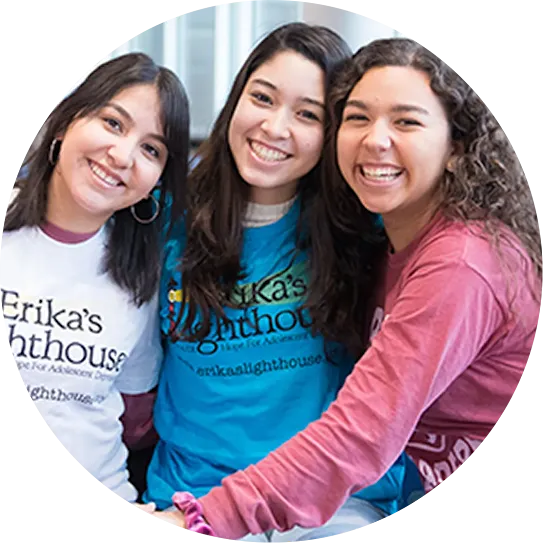 Three young women smiling and posing closely together while wearing branded "Erika's Lighthouse" t-shirts, cropped into a neat circular frame.