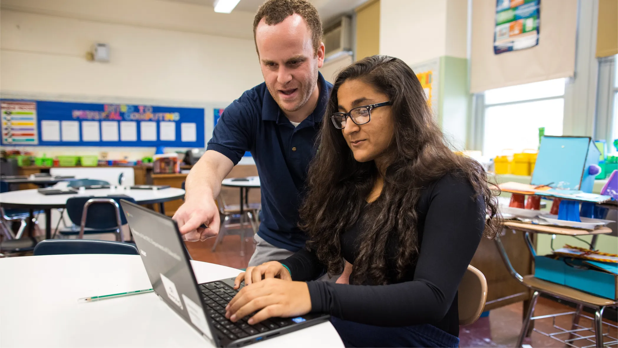 A male teacher in a navy polo shirt points to a Chromebook screen while assisting a female student with long dark hair in a bright, modern classroom setting.