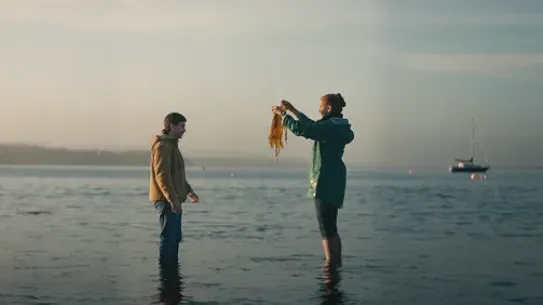 Two people stand in shallow water at the beach as one holds up seaweed; a boat floats in the distance.