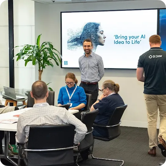 A group of professionals participating in a workshop around a conference table. A man in a striped shirt stands smiling near a large digital screen displaying the text "Bring your AI Idea to Life". Several colleagues sit at the table taking notes or collaborating, while a man in an "IX Crew" polo shirt walks past on the right.