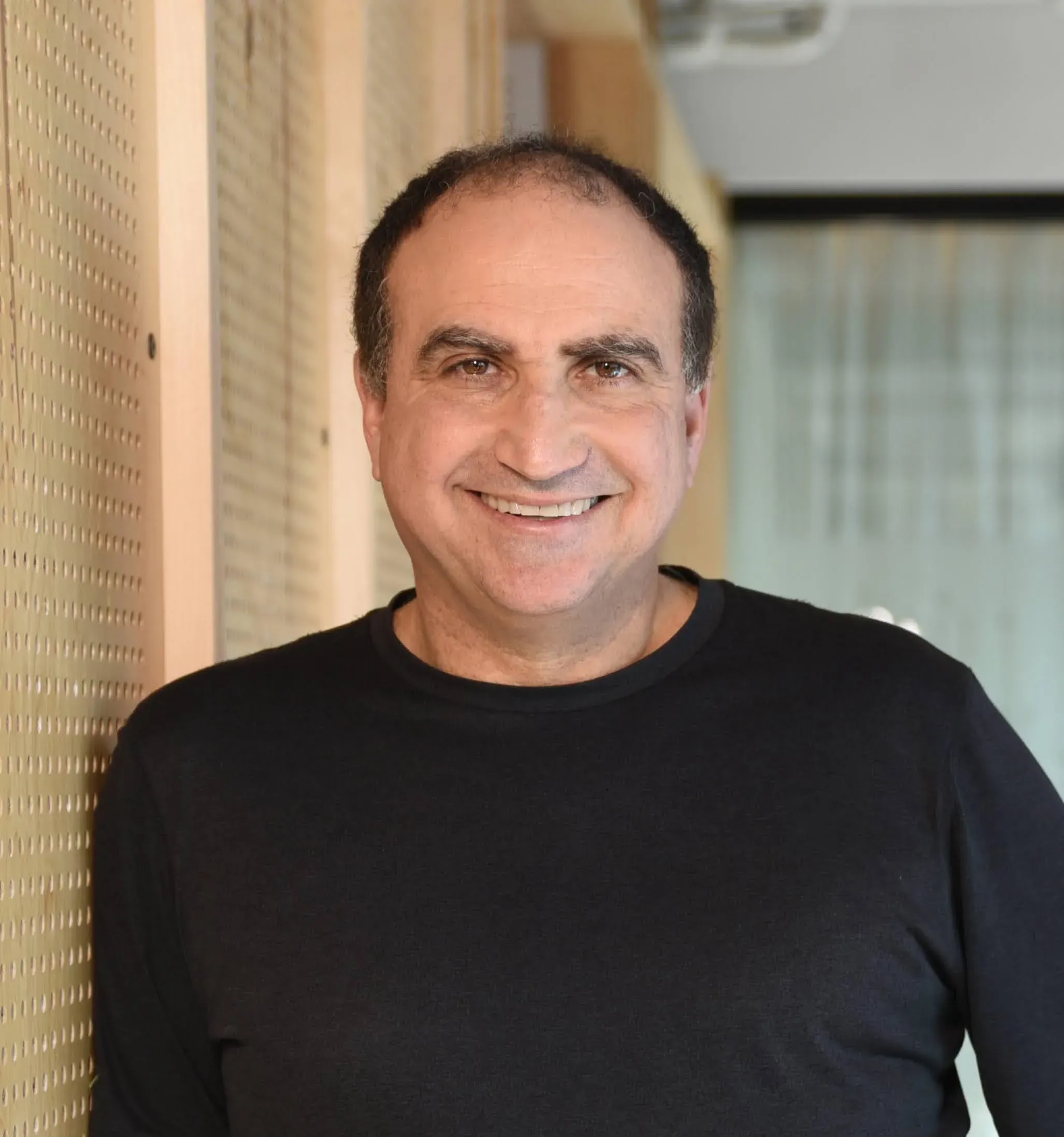 A professional headshot portrait of Yossi Matias, VP & GM of Google Research and Crisis Resilience Lead, smiling and wearing a casual black long-sleeve shirt while leaning against a textured, light-colored wooden wall.