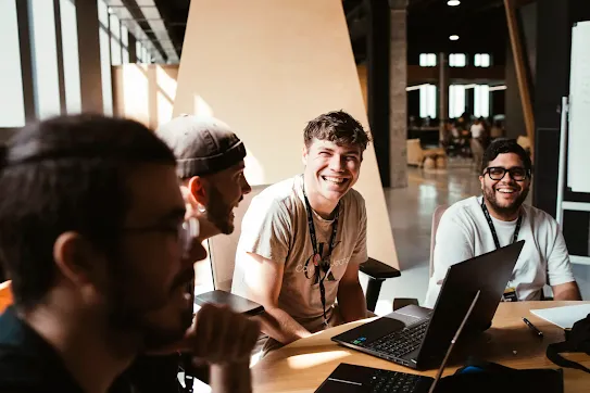 Four young men sitting around a table with laptops, laughing and working together in a modern workspace.