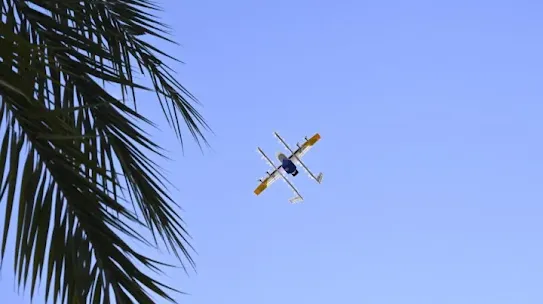A white drone with yellow and blue accents flying in a clear, bright blue sky