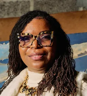 A smiling Black woman with long locs, wearing stylish tortoiseshell cat-eye glasses, a white vest, and a large gold necklace, posing for a headshot against a rustic background.